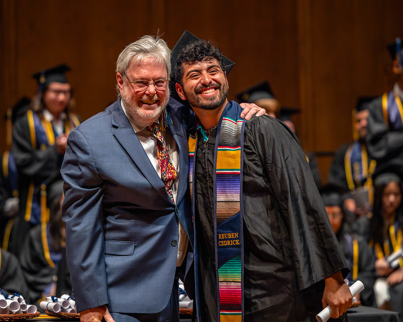 Man in blue suit stands with student in graduation regalia