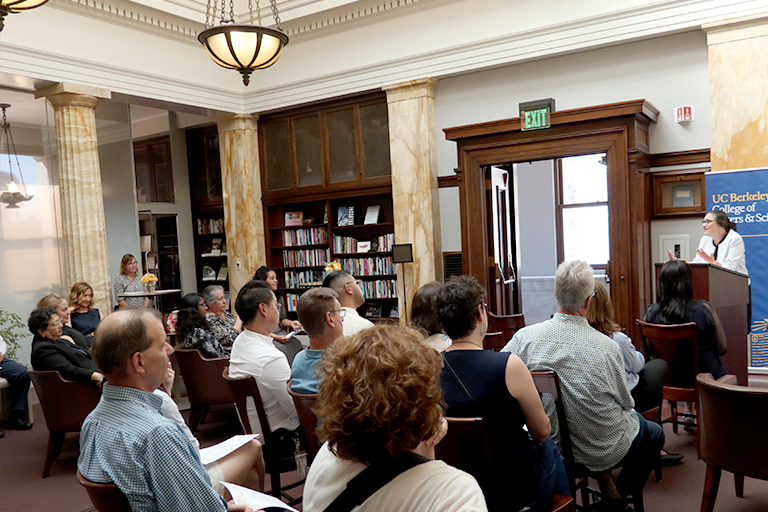 Guests sit indoors for a ceremony, listening to a woman address them at a lectern. 