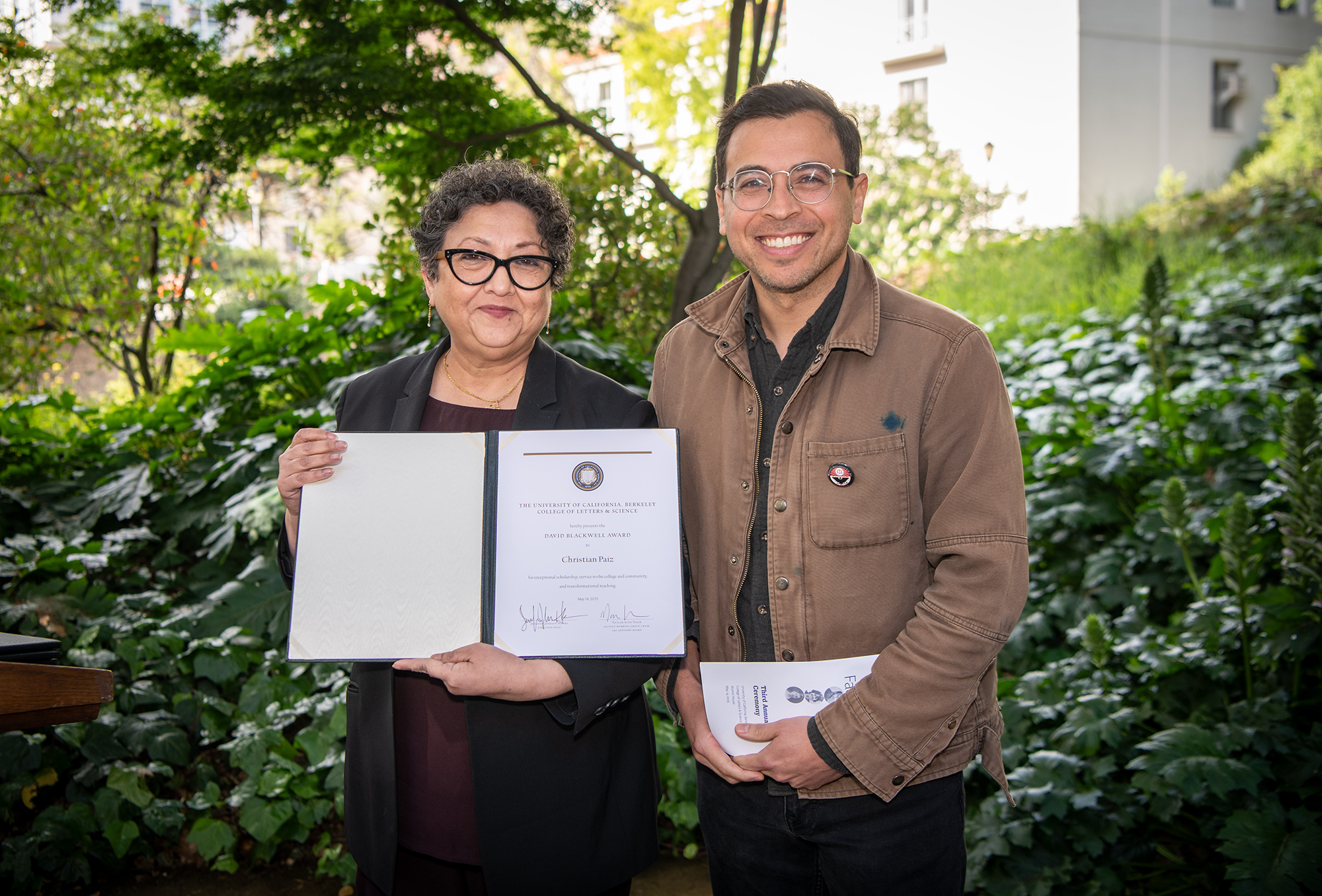 Two people stand outside, holding up award certificates