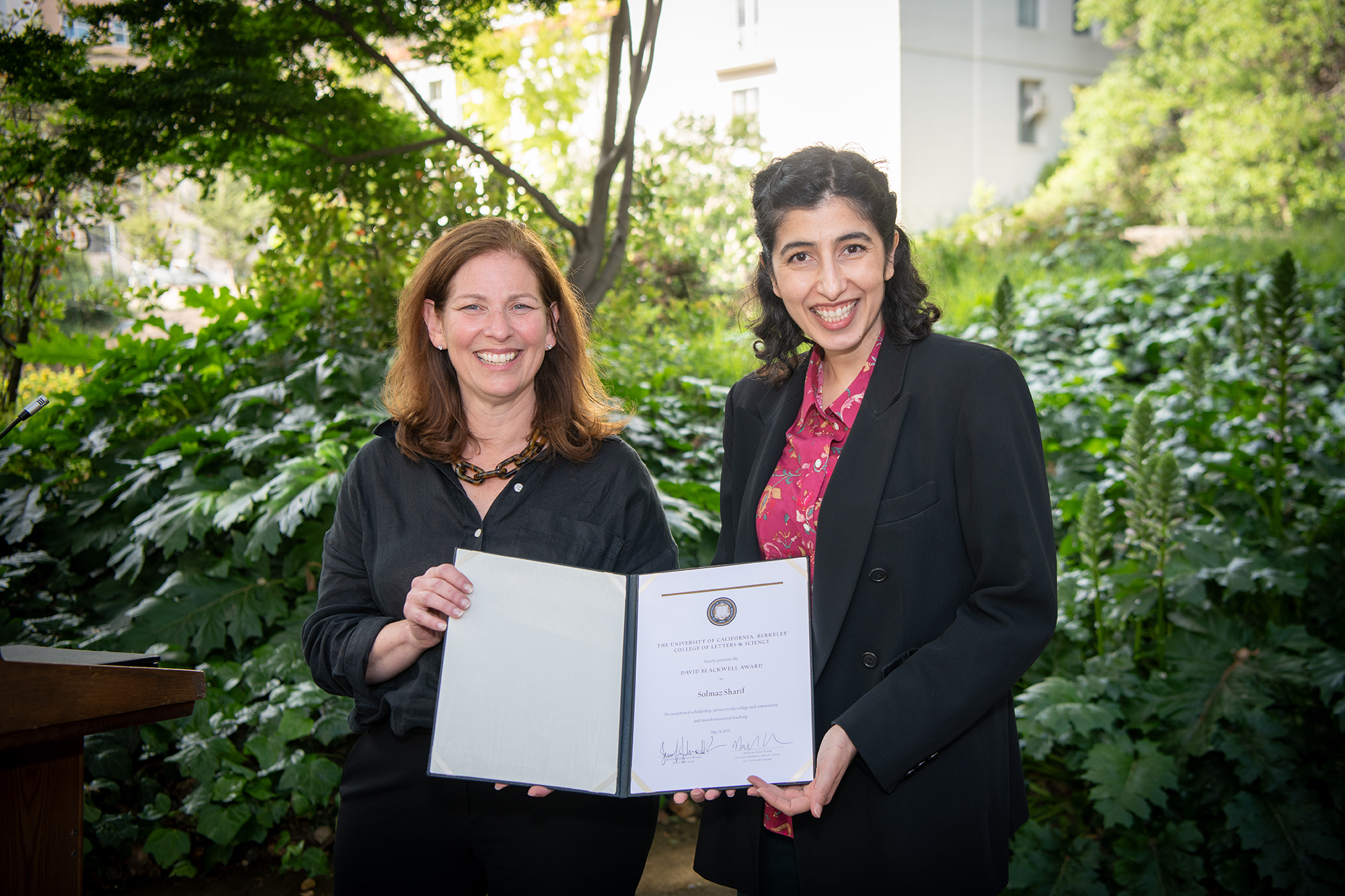 Two people stand outside, holding up award certificates