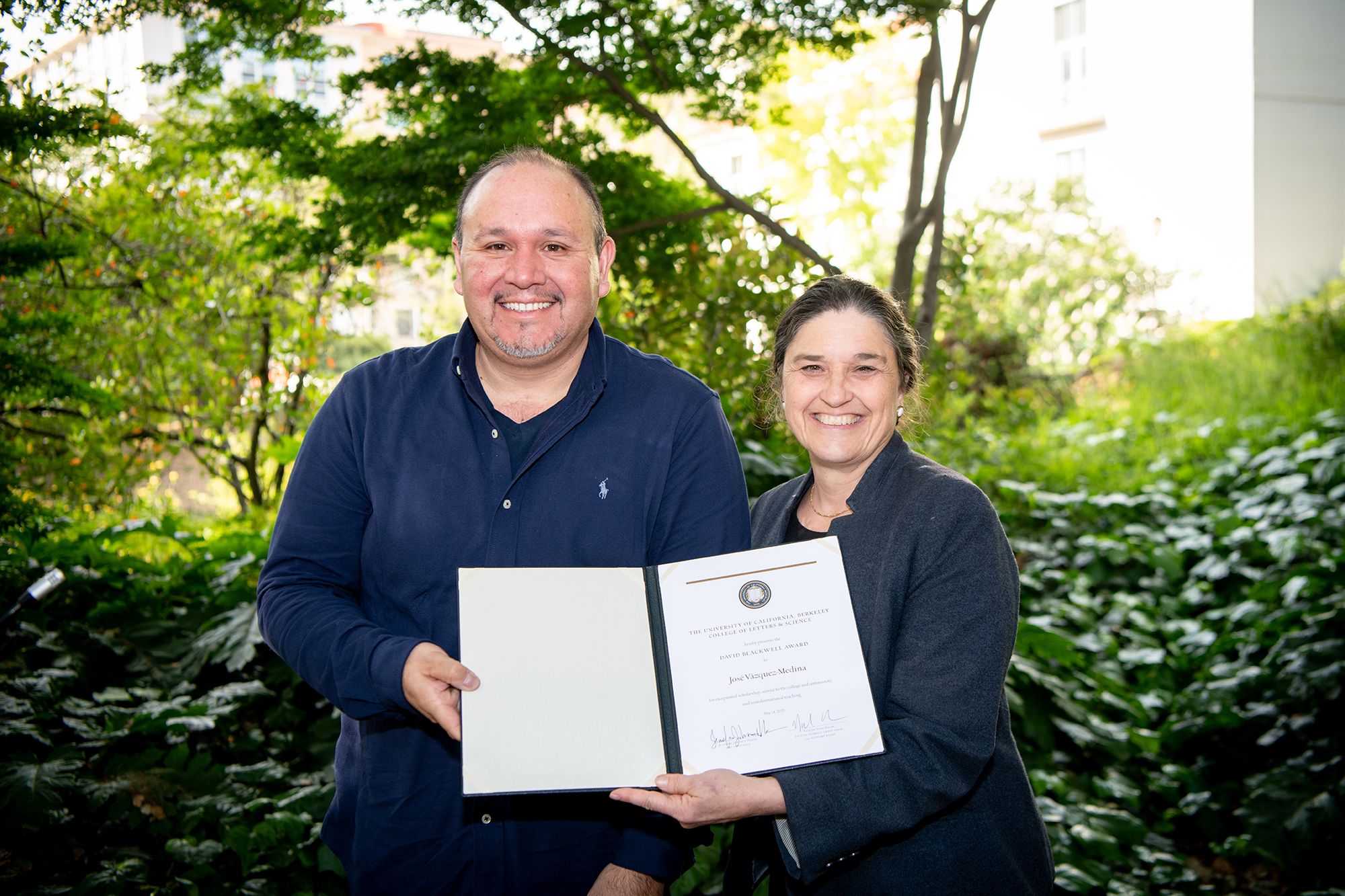 Two people stand outside, holding up award certificates