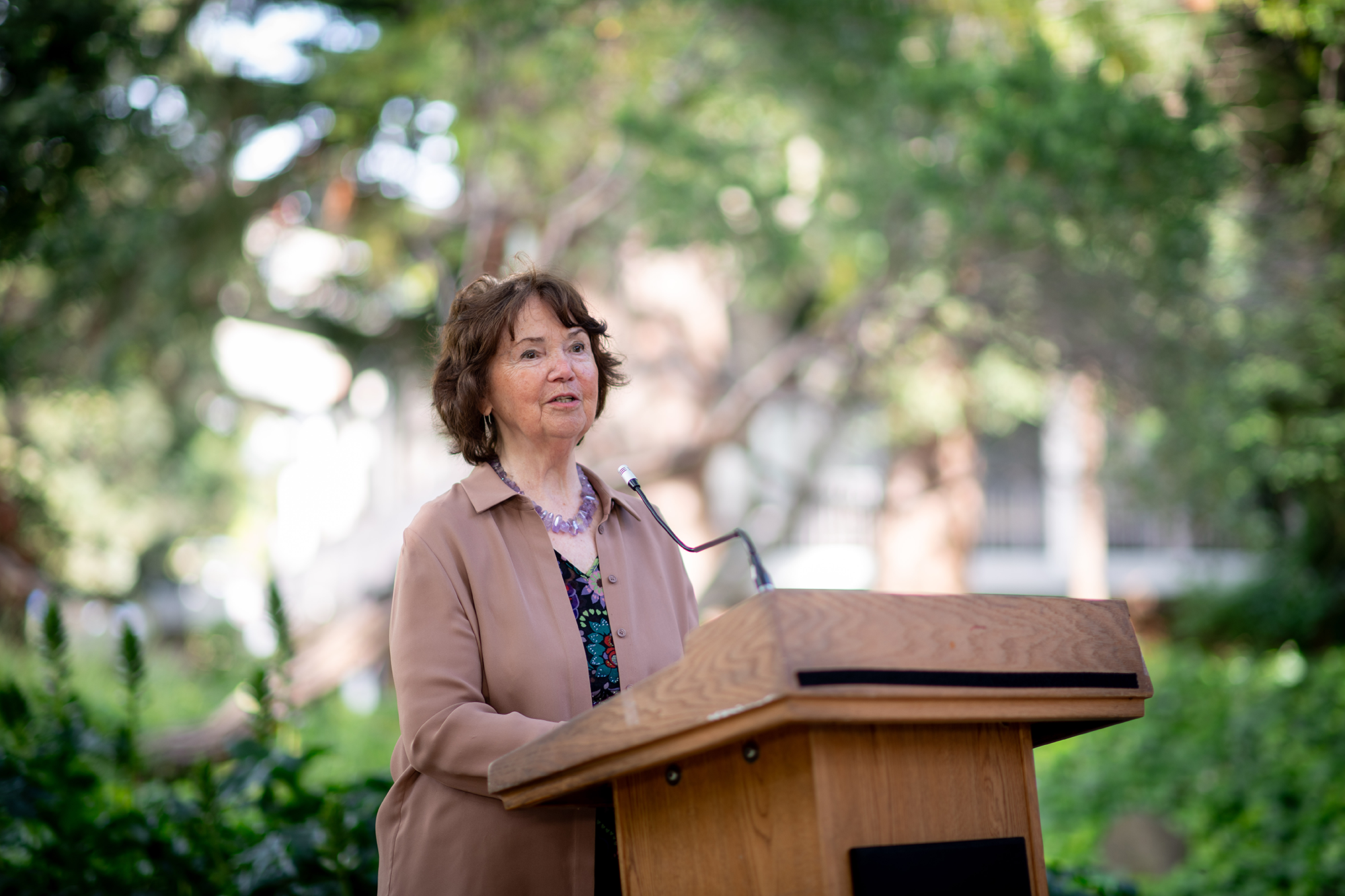 Woman stands at a lecturn speaking to an audience outdoors