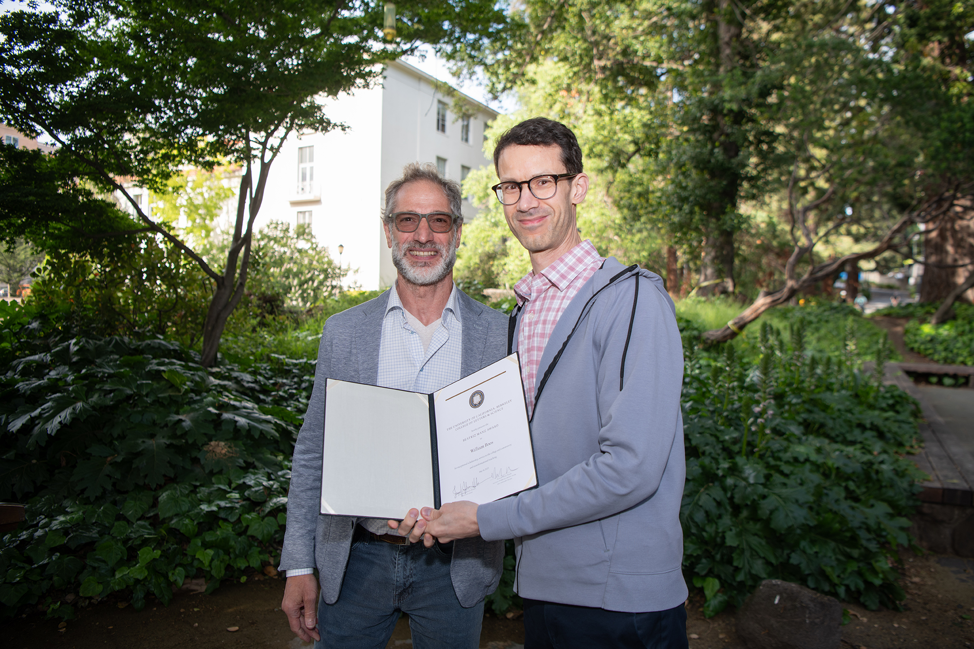 Two people stand outside, holding up an award certificate