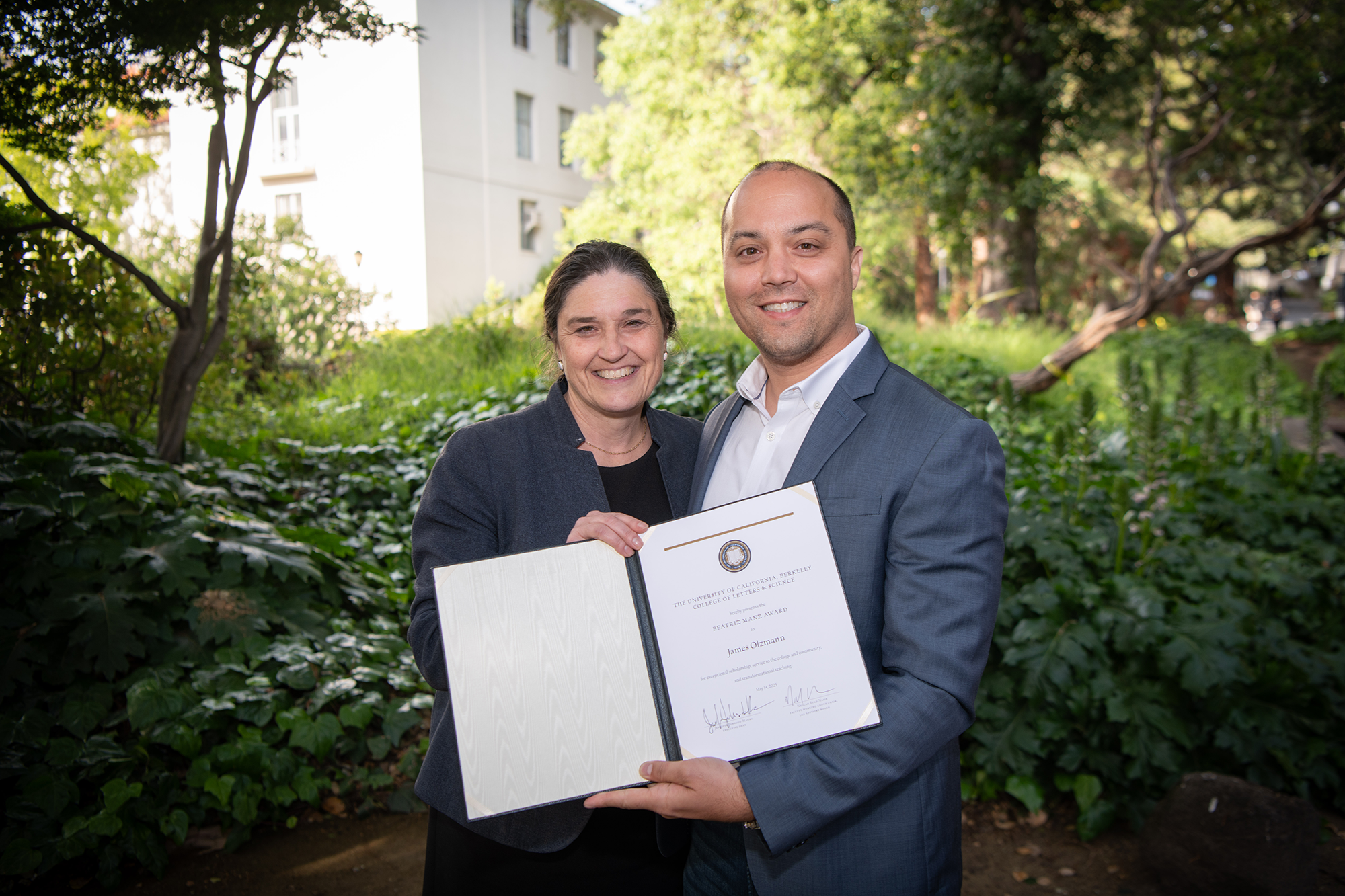 Two people stand outside, holding up an award certificate