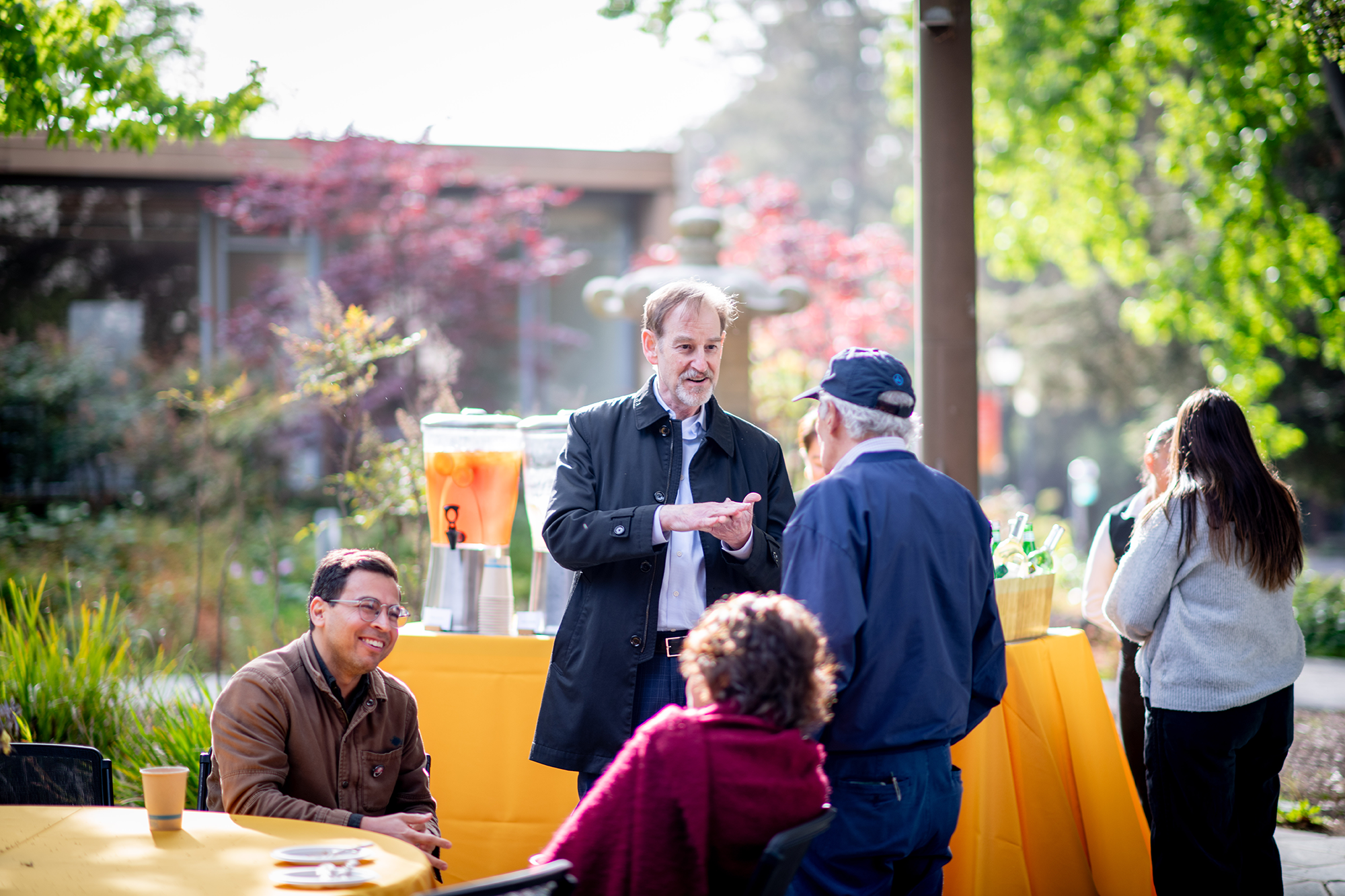 Man at an outdoor reception gestures while speaking to someone, while guests chat nearby