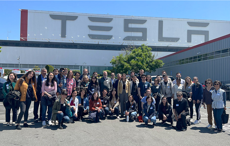A group of over 40 people stand in front of a large gray industrial building with "TESLA" written on top.