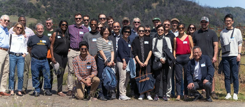 Dozens of people pose for a group photo on a sunny day with wooded hills in the background.