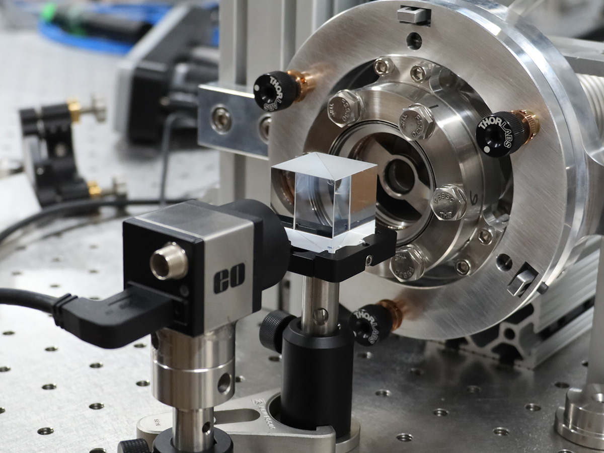 A device that emits laser beams is pointed at a cube in front of a metal chamber