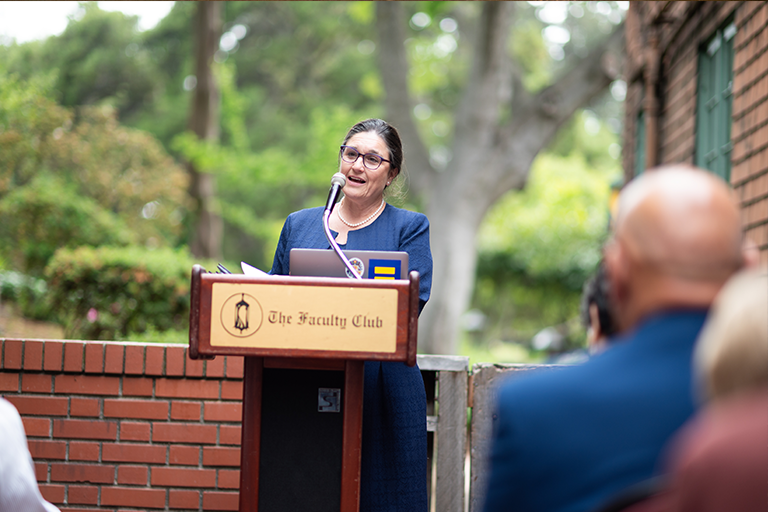 Woman stands at podium and addresses an audience outdoors