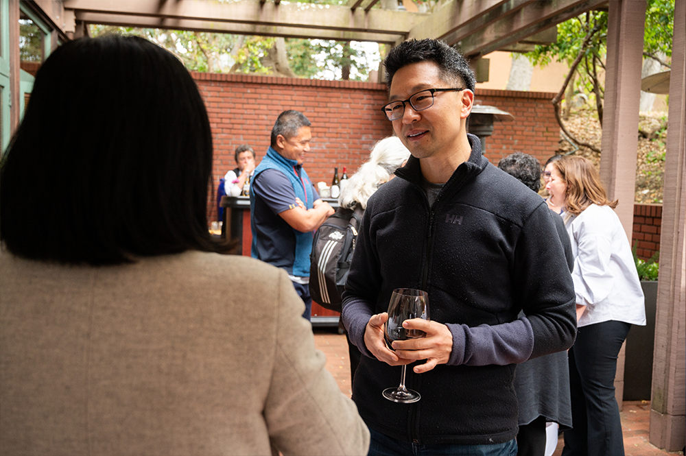 Man with dark hair and glasses wearing a dark pullover holds a glass of wine and chats 