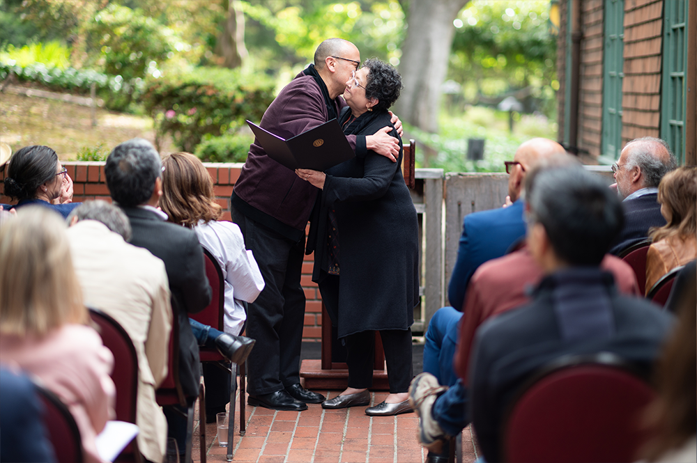 Two people in front of a podium hug as they hold an award