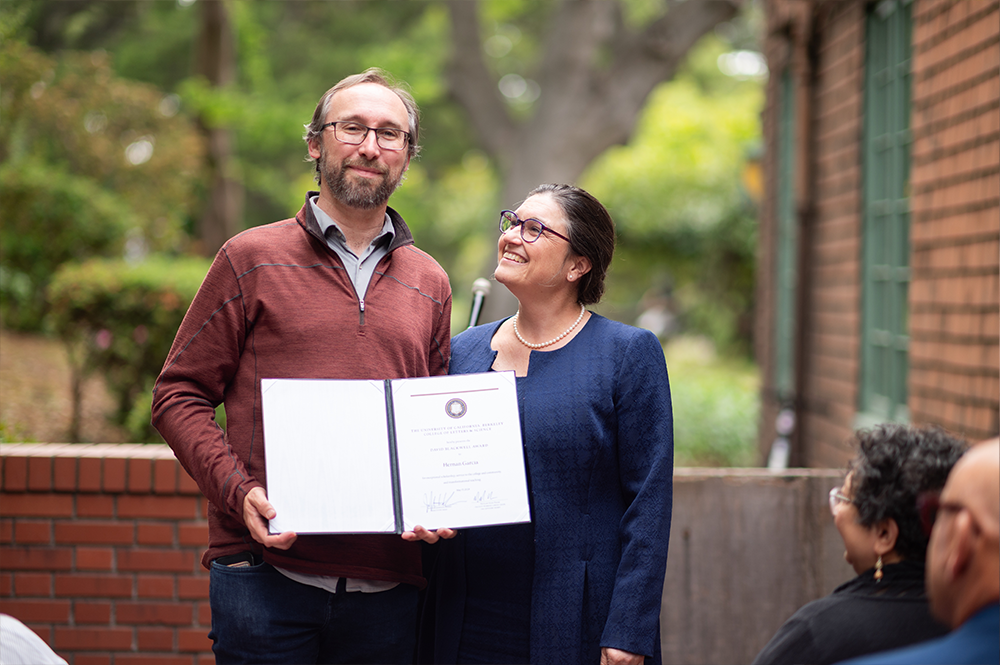 Man with brown sweater holds his award while standing next to a woman with blue suit