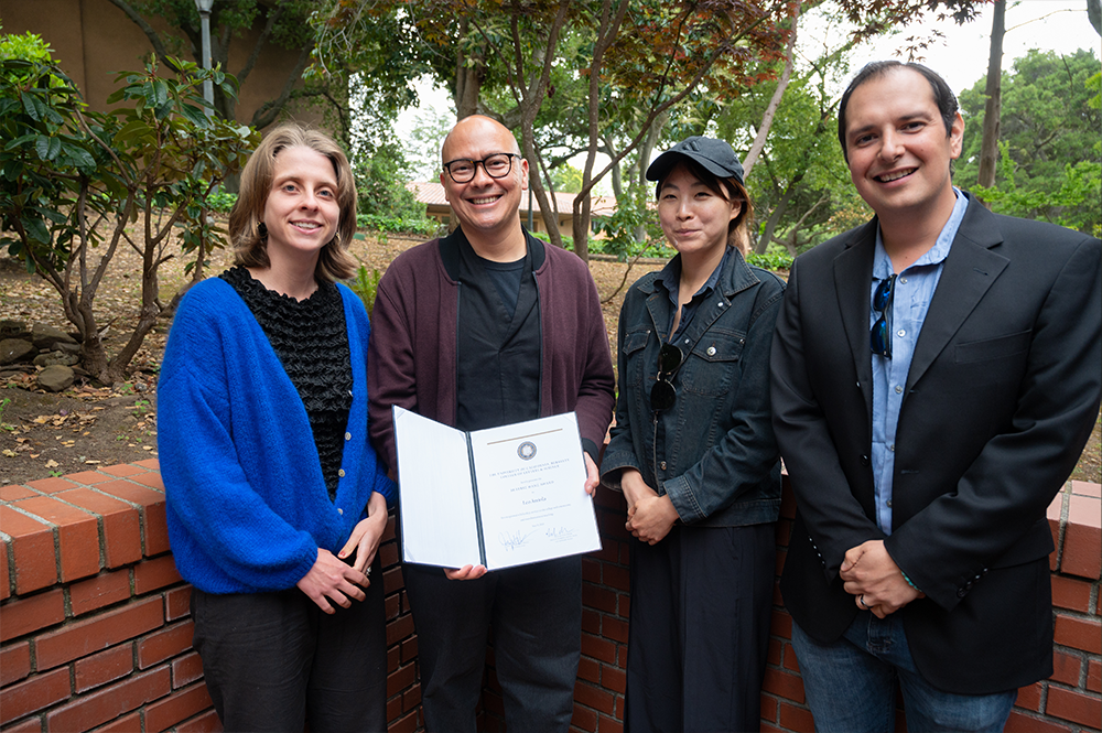 Man wearing purple jacket and dark shirt holds an award, surrounded by three guests