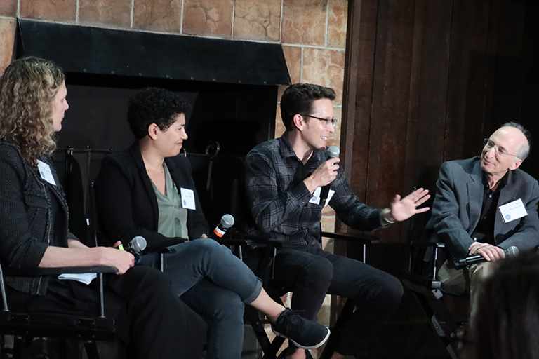 Three panelists watch a fourth panelist speak animatedly, gesturing with his hands