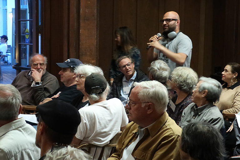 A man holds a mic in an audience, poised to ask a question to a panel