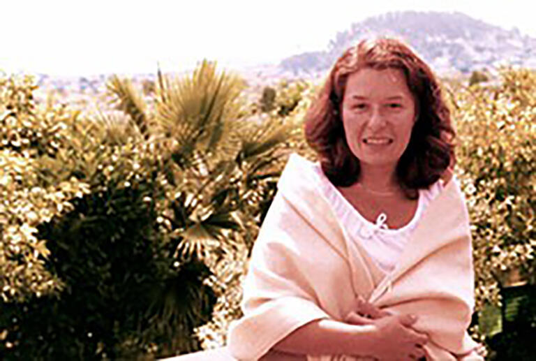 A woman in a light-colored wrap looks at the camera with trees in the background.