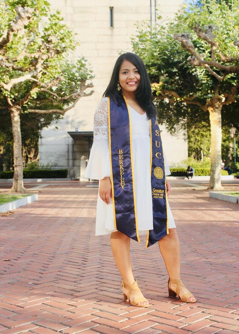 Woman with long dark hair wearing a dress and graduation regalia, smiling at camera