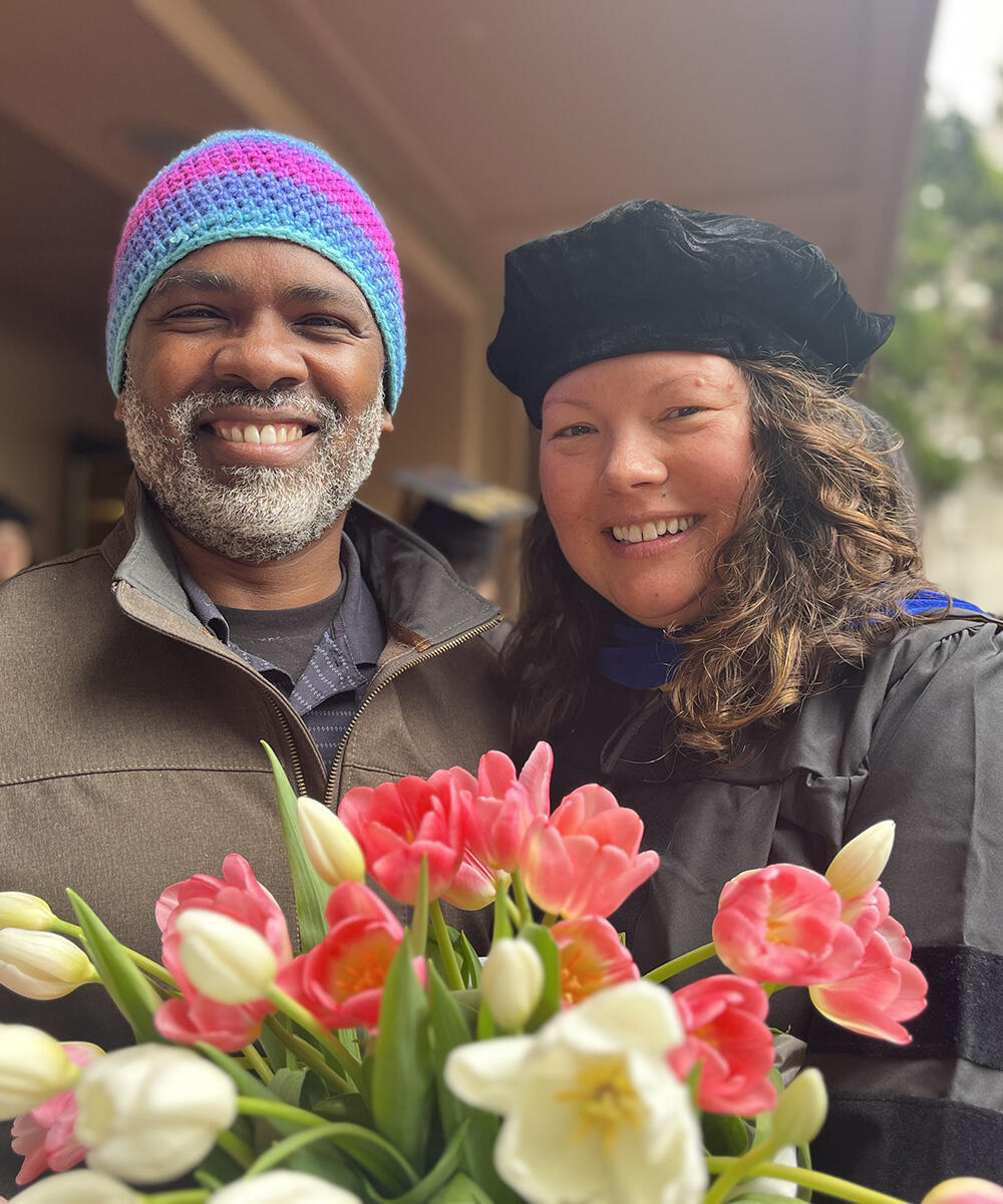 A man in a colorful hat and a woman in graduation attire smile behind flowers