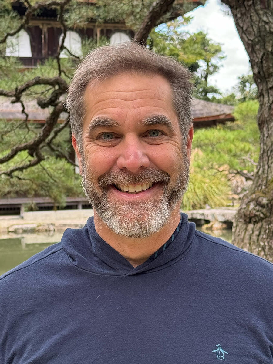 A bearded man smiles in front of an interesting tree and building