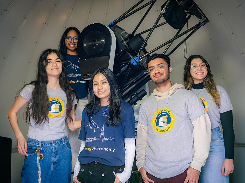 Five students wearing Department of Astronomy clothing pose next to a telescope that is several feet long.