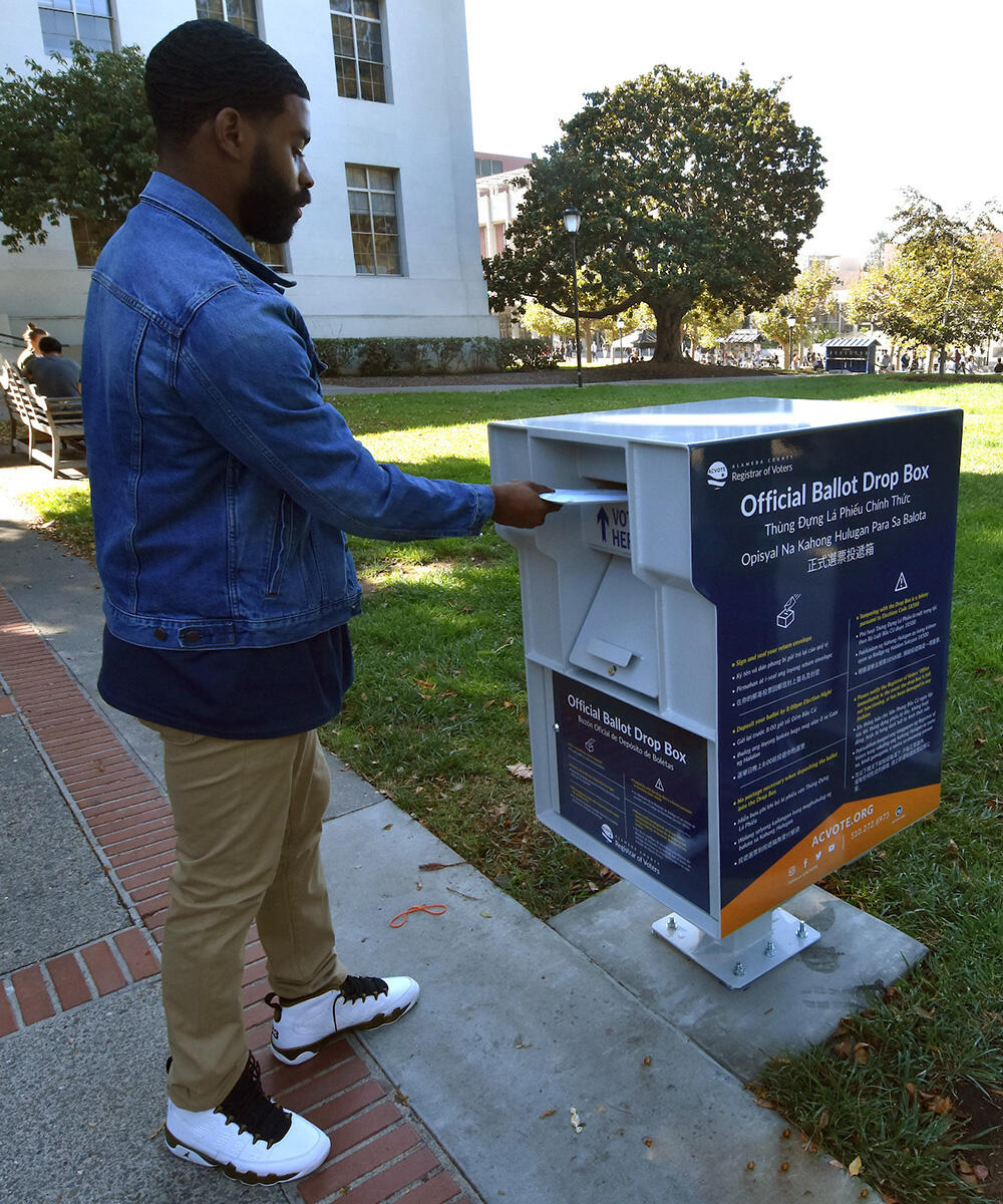 A voter wearing a denim jacket slides an envelope into a ballot drop box.