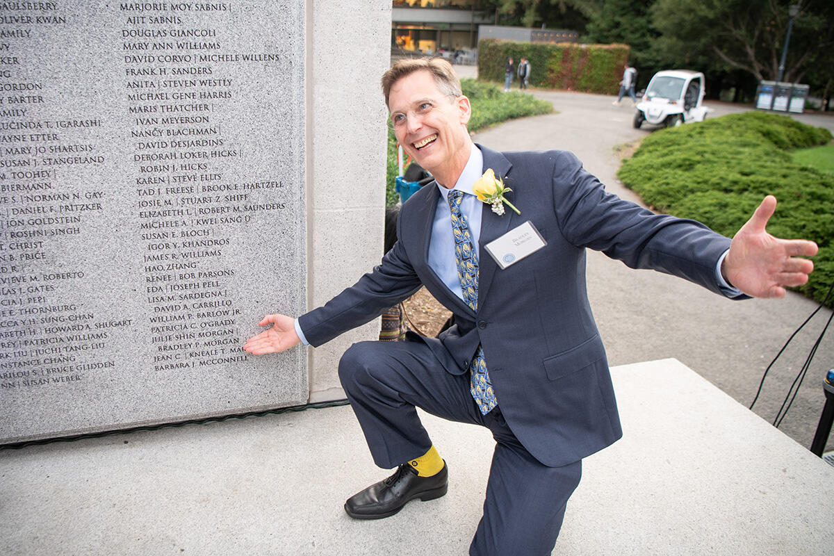 Brad Morgan, on one knee, smiles and gestures toward his name on the Builders of Berkeley wall