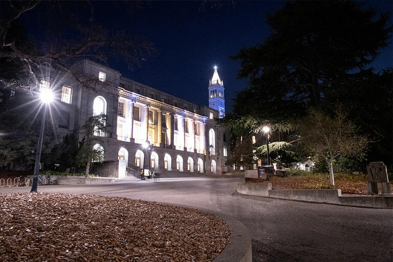 White building and tower on a campus lit up at night, with a street lamp glowing in the dark