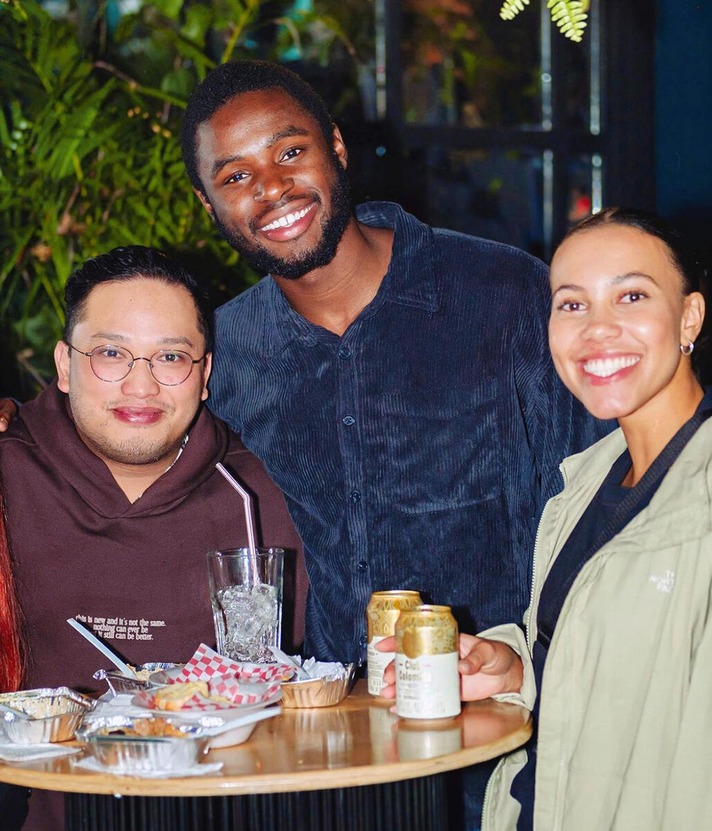 Chaka stands next to two friends with food and drinks at a tall table