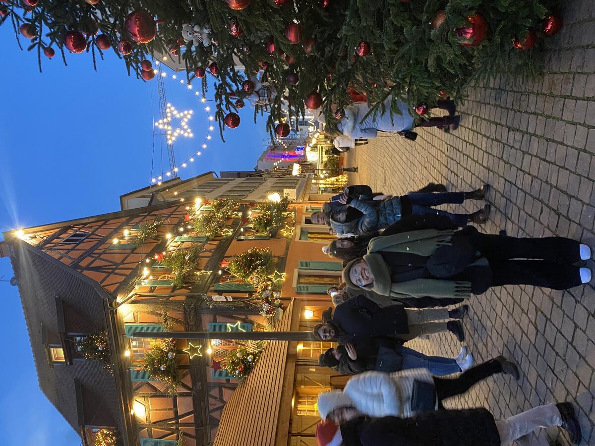 Woman wearing a scarf smiles while standing outside in a holiday market at night, next to a Christmas tree and a house decorated with holiday lights