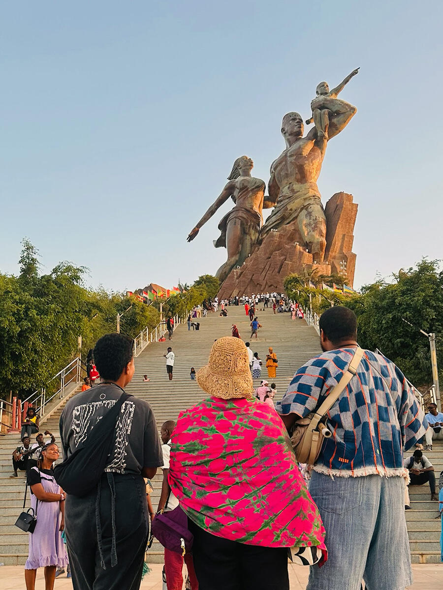 Three people look up a long series of steps at a statue of a man holding a baby and a woman