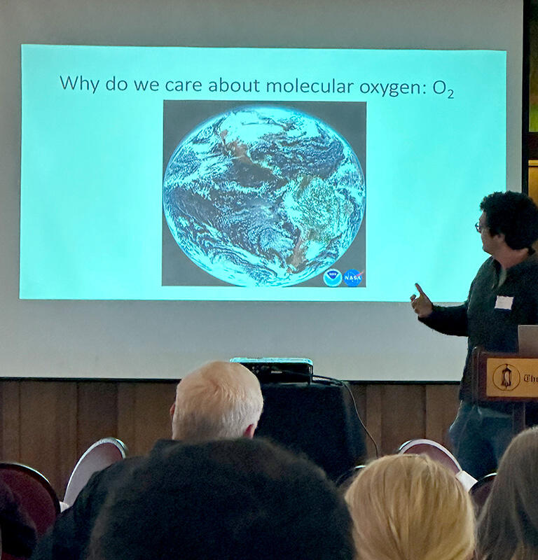 Man standing behind a lecturn turned to a presentation on a screen behind him, which has an image of Earth and the question, Why do we care about molecular oxygen?