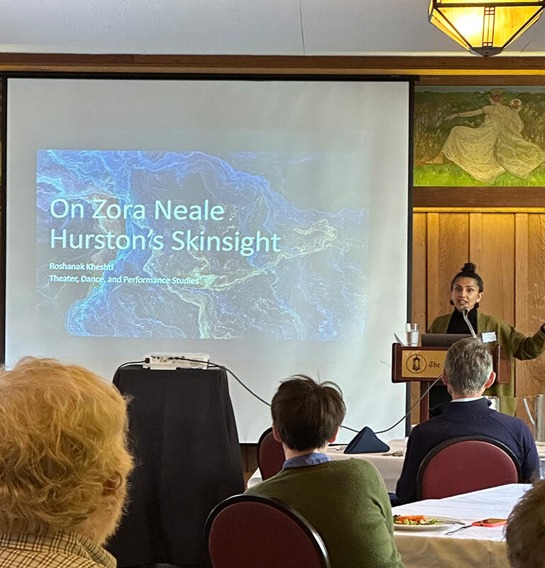 Woman stands at a lecturn speaking to an audience. Presentation behind her reads, "On Zora Neale Hurston's Skinsight"