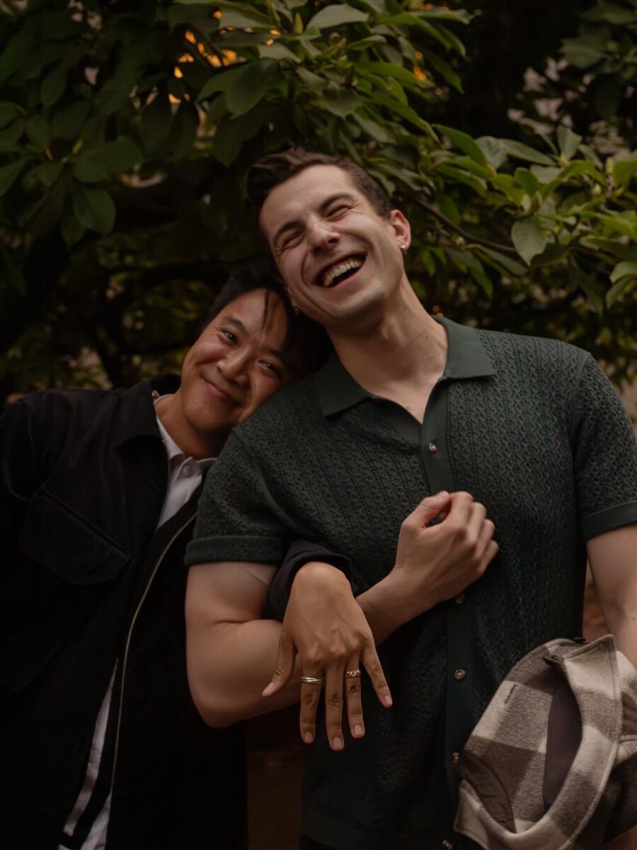 A newly engaged couple smile at camera: one man holds up his ring fingers to show engagement and the other man throws head back in laughter