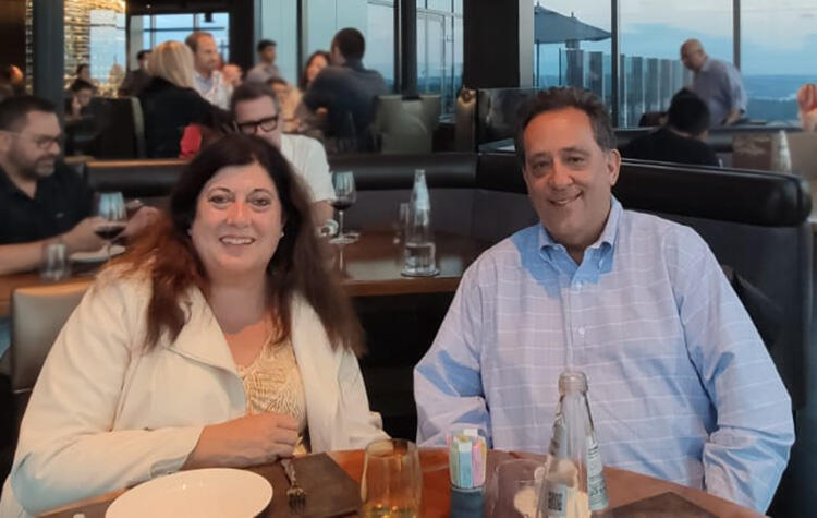 Mike and Melanie smiling at a restaurant table with other patrons and large windows in the background