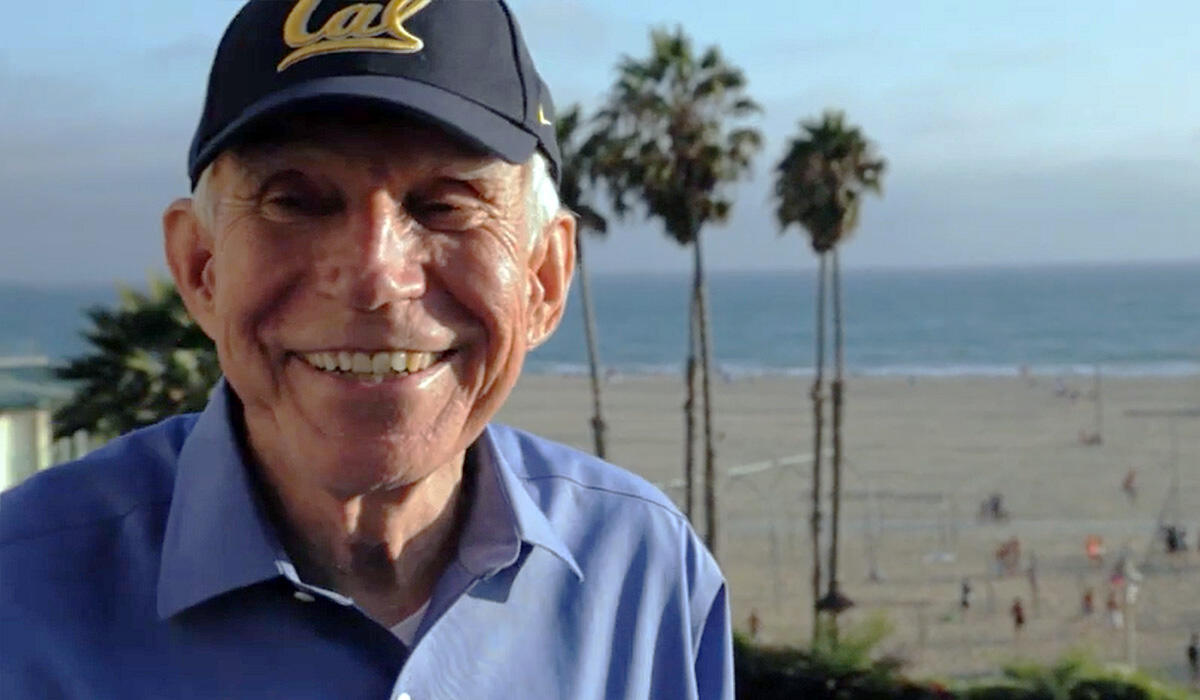 A man in a Cal hat smiles in front of a Southern California beach and some palm trees