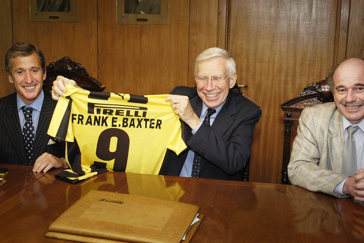 Three smiling men sit at a table. The one in the middle holds up a bright Pirelli jersey with the name Frank E. Baxter and the number 9.