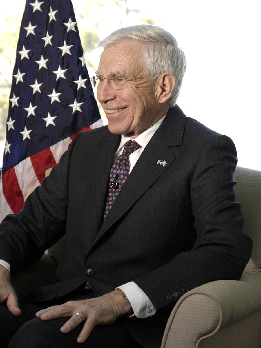 A man in a suit sits in a chair in front of an American flag
