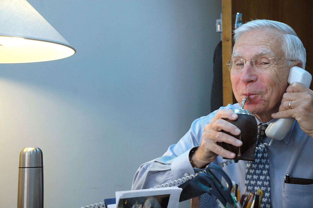 A man with a wry smile looks at the camera while sipping from a straw and holding a phone to his ear