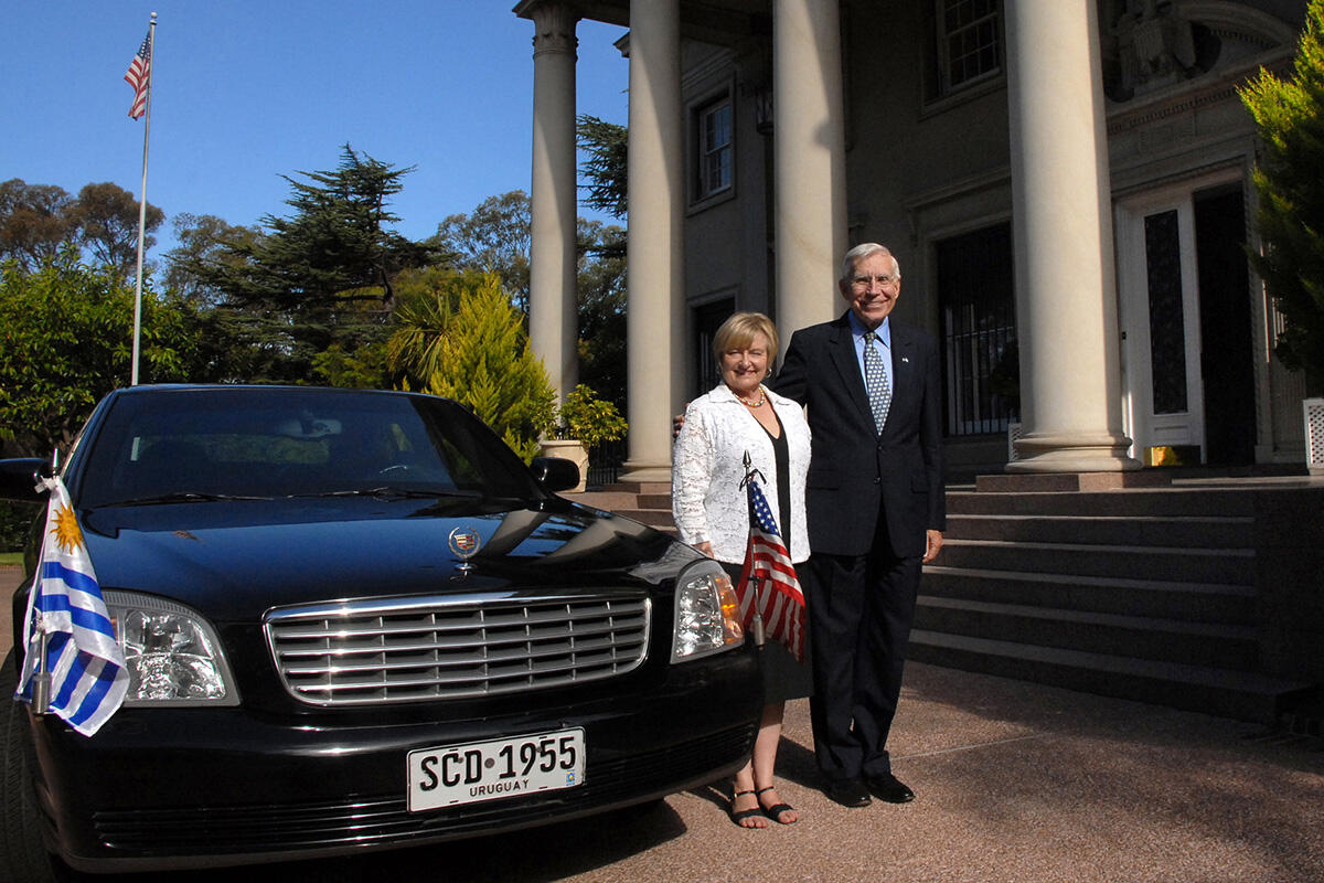 A man and woman stand next to a dark diplomatic vehicle and a house fronted by large columns