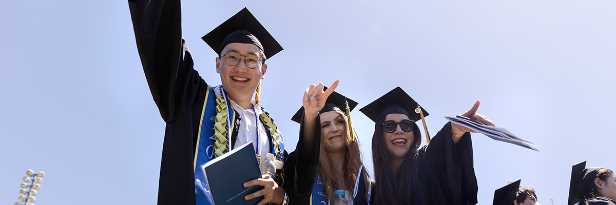 Three graduates smile and point at camera, wearing their regalia from outside, blue sky behind them