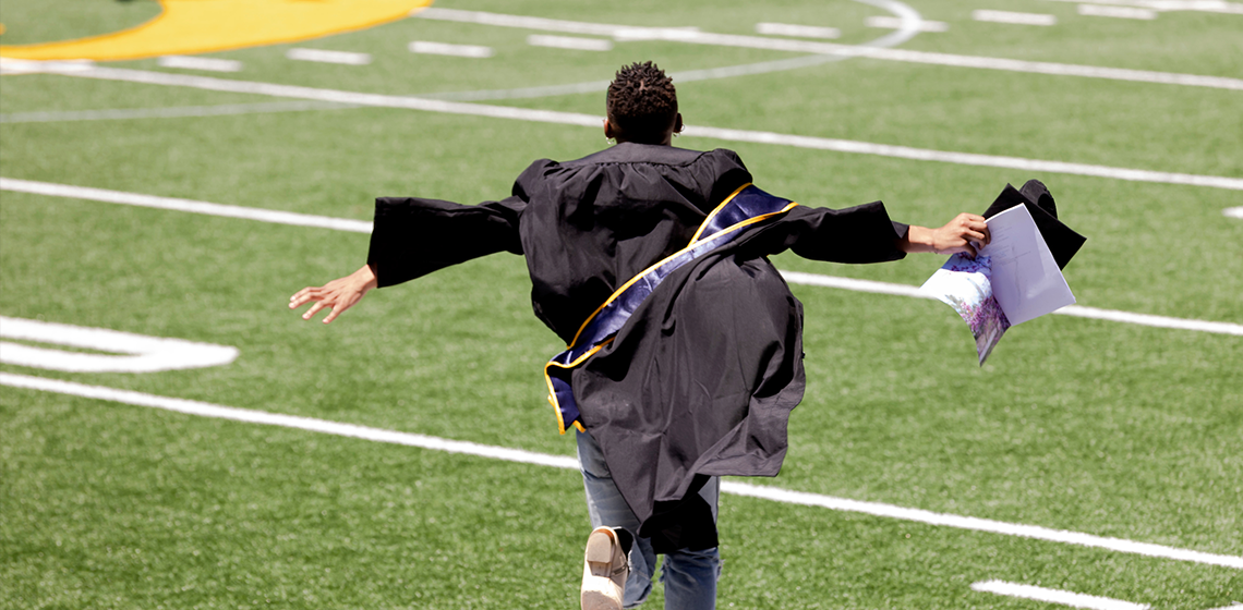 Graduate in a gown runs across a field with arms outspread, holding a diploma and cap in one hand