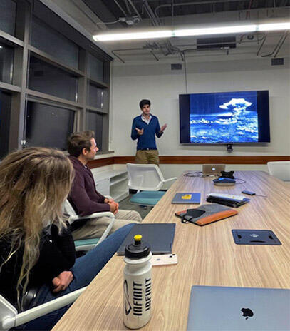 Two students sit at a table listening to another person give a presentation by a large screen.