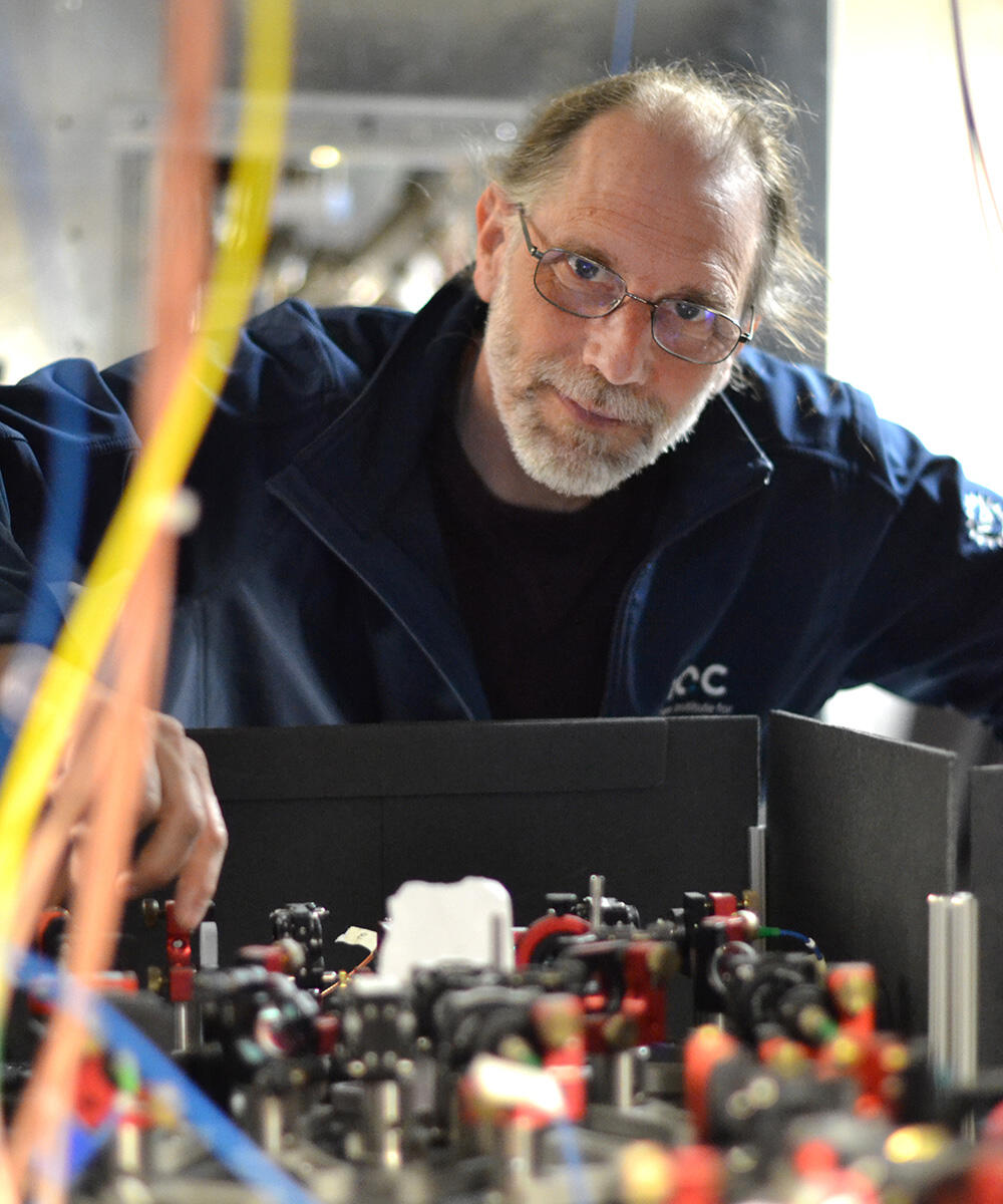 A man with glasses and a blue jacket tinkers with equipment while looking at the camera