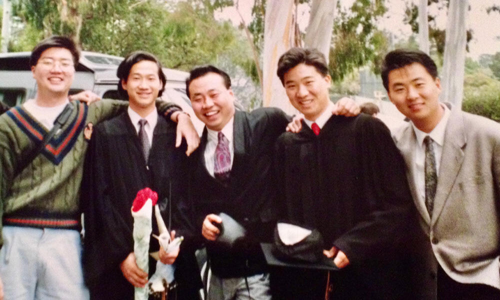 Five college-aged men smile for a photo. Two are wearing graduation robes.