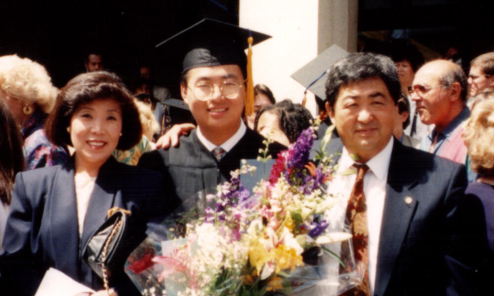 A man in black graduation regalia surrounded by his parents in suit jackets