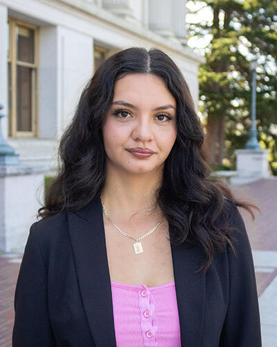 A woman wears a pink shirt, black suit jacket, and necklace on the UC Berkeley campus