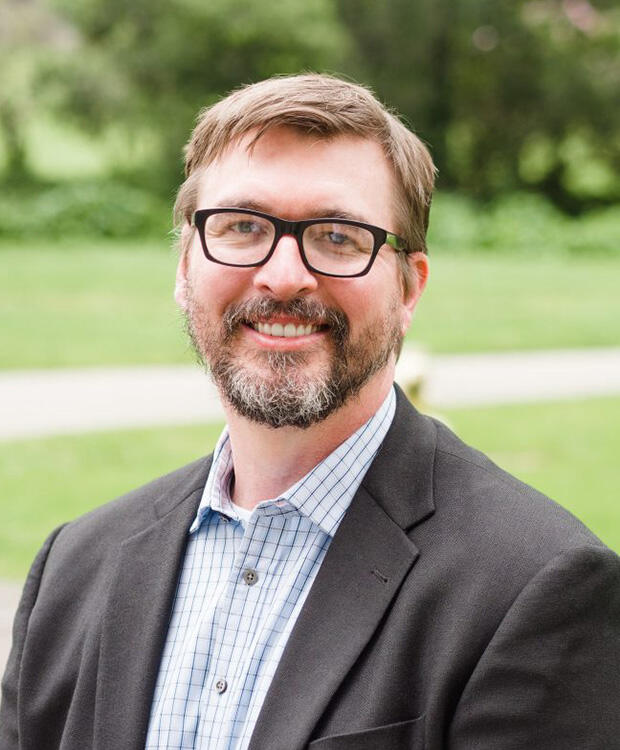 A smiling man wearing glasses, a suit jacket, and a patterned shirt