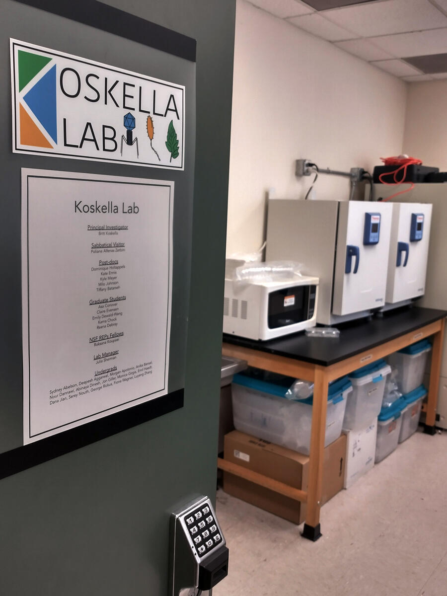 Signs on an open door mark the room as the Koskella Lab, with lab equipment in the background.