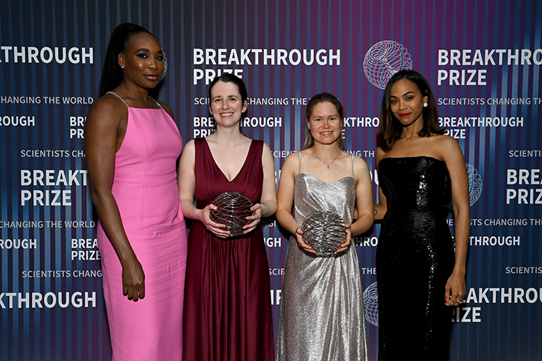 Four women in formal dresses in front of Breakthrough Prize step and repeat.