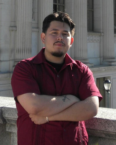 A man in a red shirt crosses his arms in front of a UC Berkeley building